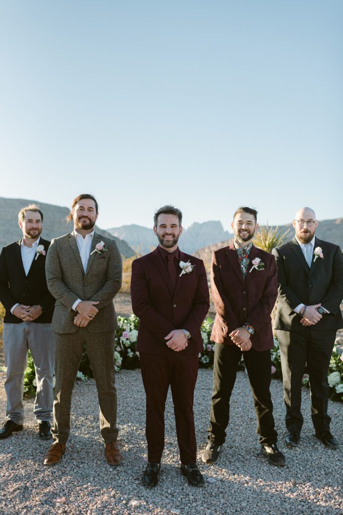 groom posing with four groomsman in V shape with mismatching suits groom in Burgundy suit and tie