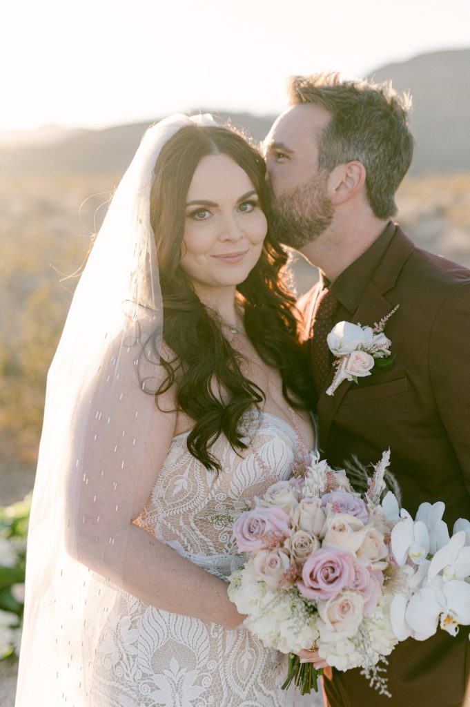 Groom softly kissing bride wearing veil and lace wedding gown in red rock canyon bride is holding a white and pink rose bouquet with white orchids