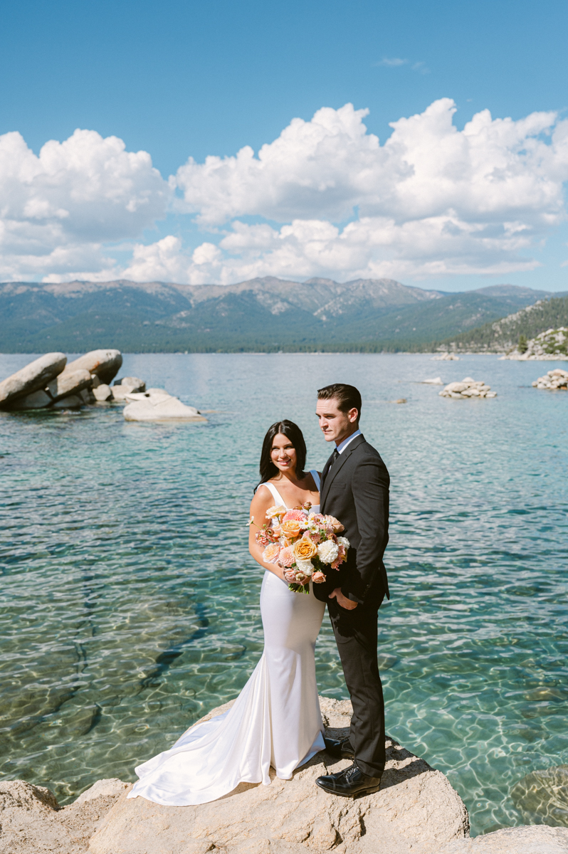 bride and groom standing on white boulder over blue waters in Lake Tahoe